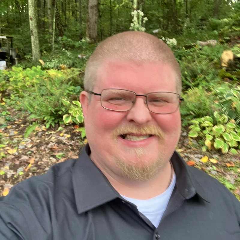 A close-up selfie of a smiling blond man with glasses, a mustache, and goatee, wearing a black shirt in a lush, green forest setting.